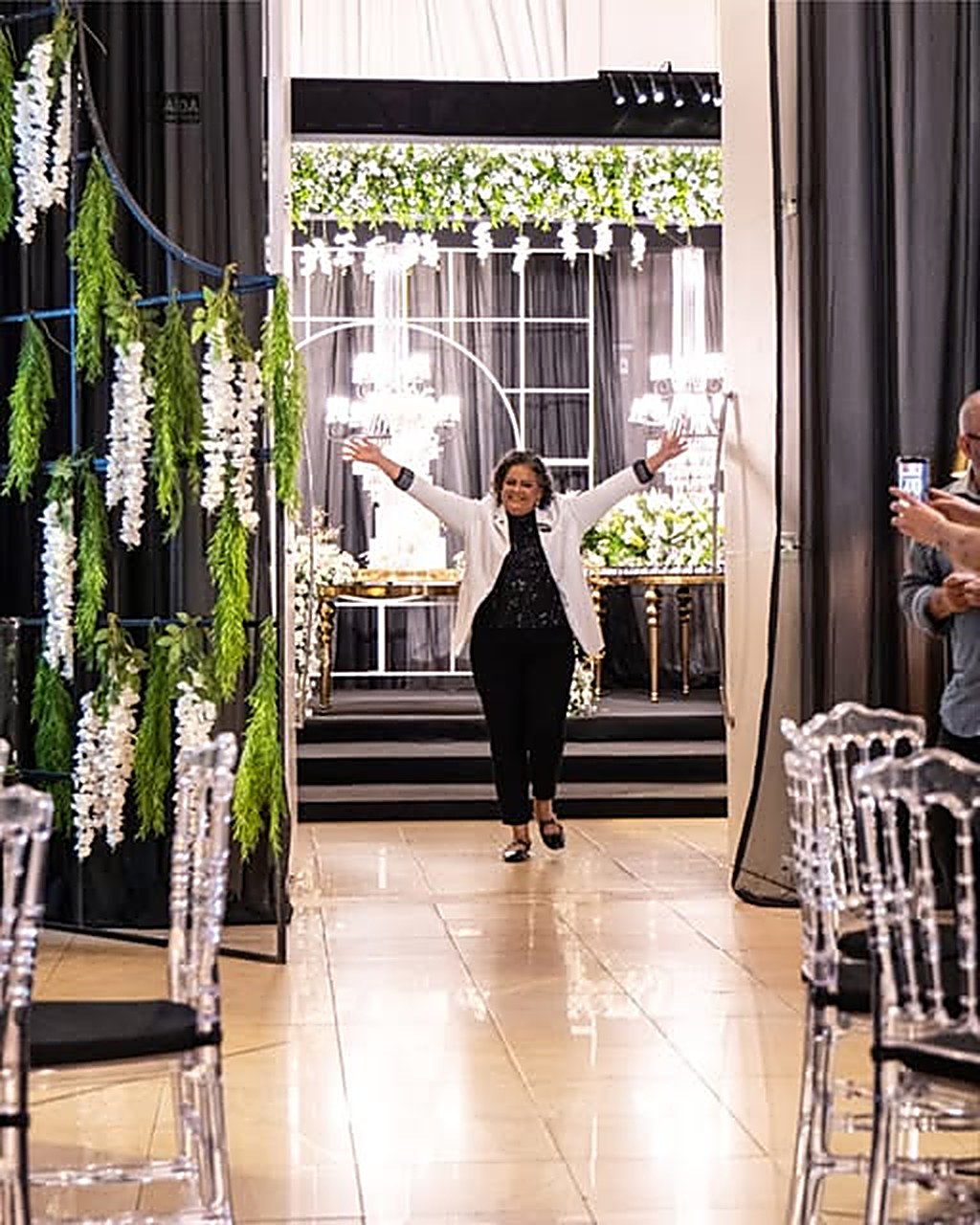 Salão de festas decorado com cortinas de folhagens, lustres de cristal e cadeiras transparentes